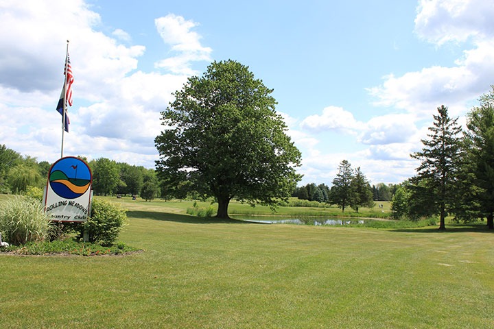 Rolling Meadows sign under the US flag, with trees and lake in the background.